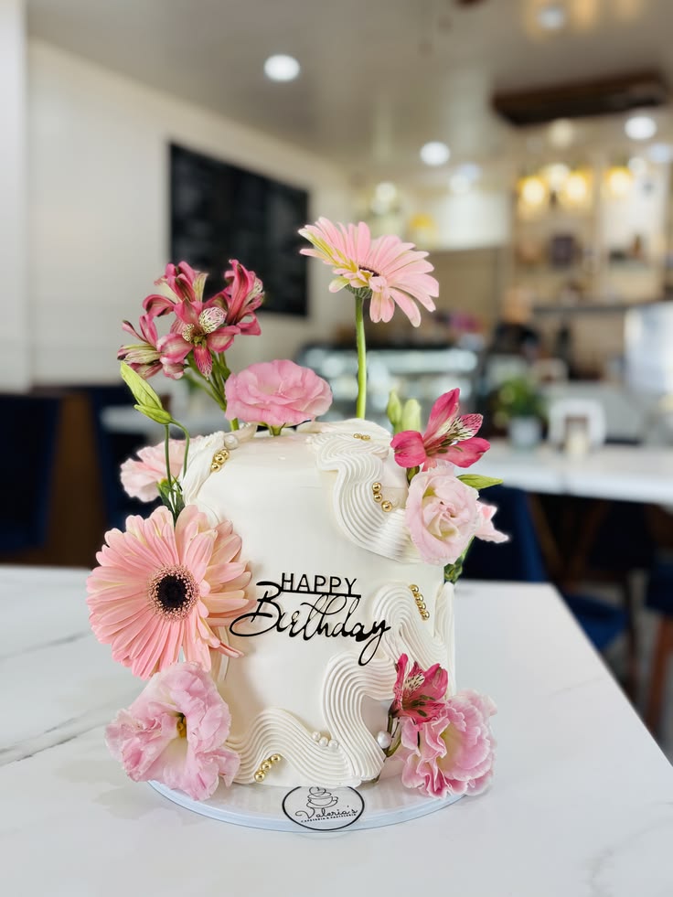 Un pastel blanco decorado con gerberas rosadas y más flores rosas. Los pasteles decorados con flores están en tendencia en Puebla. En la parte delantera del pastel se coloca un topper que dice "HAPPY BIRTHDAY". El pastel se coloca sobre una superficie blanca.