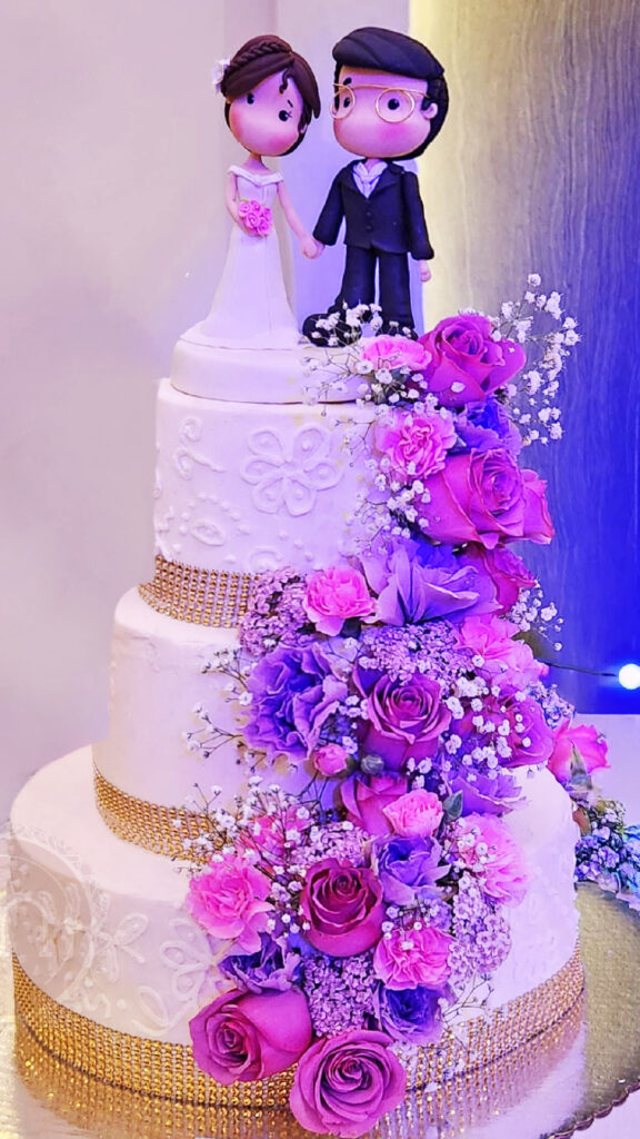 Los pasteles decorados con flores para bodas en Puebla se decoran con ramos pequeños. Este pastel tiene un acabado con textura y cintas doradas, es de tres pisos e lleva una deoración de cascada de flores moradas en diagonal. En l parte superior del pastel se colocan un par de muñecos de migajón de novios.