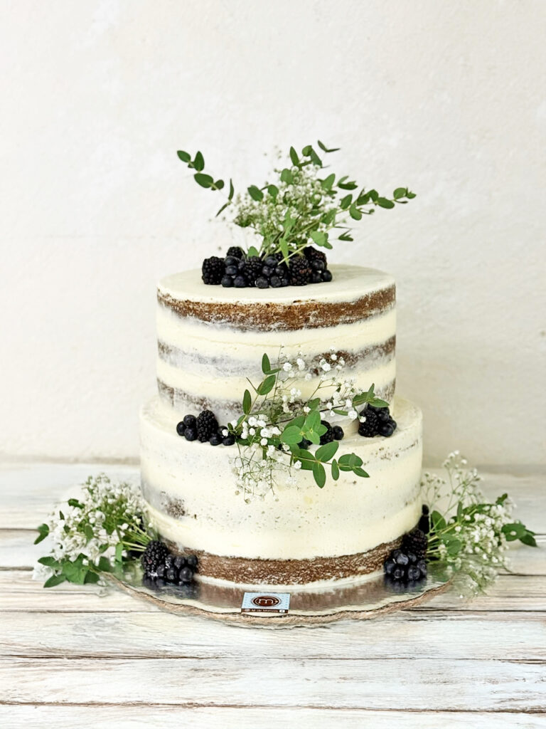 Un pastel de boda estilo naked cake de dos pisos decorado con flores y frutas como zarzamoras y arándanos. Los pasteles decorados con flores para bodas tienen diseños minimalistas. El pastel se coloca sobre una mesa de madera color blanco.